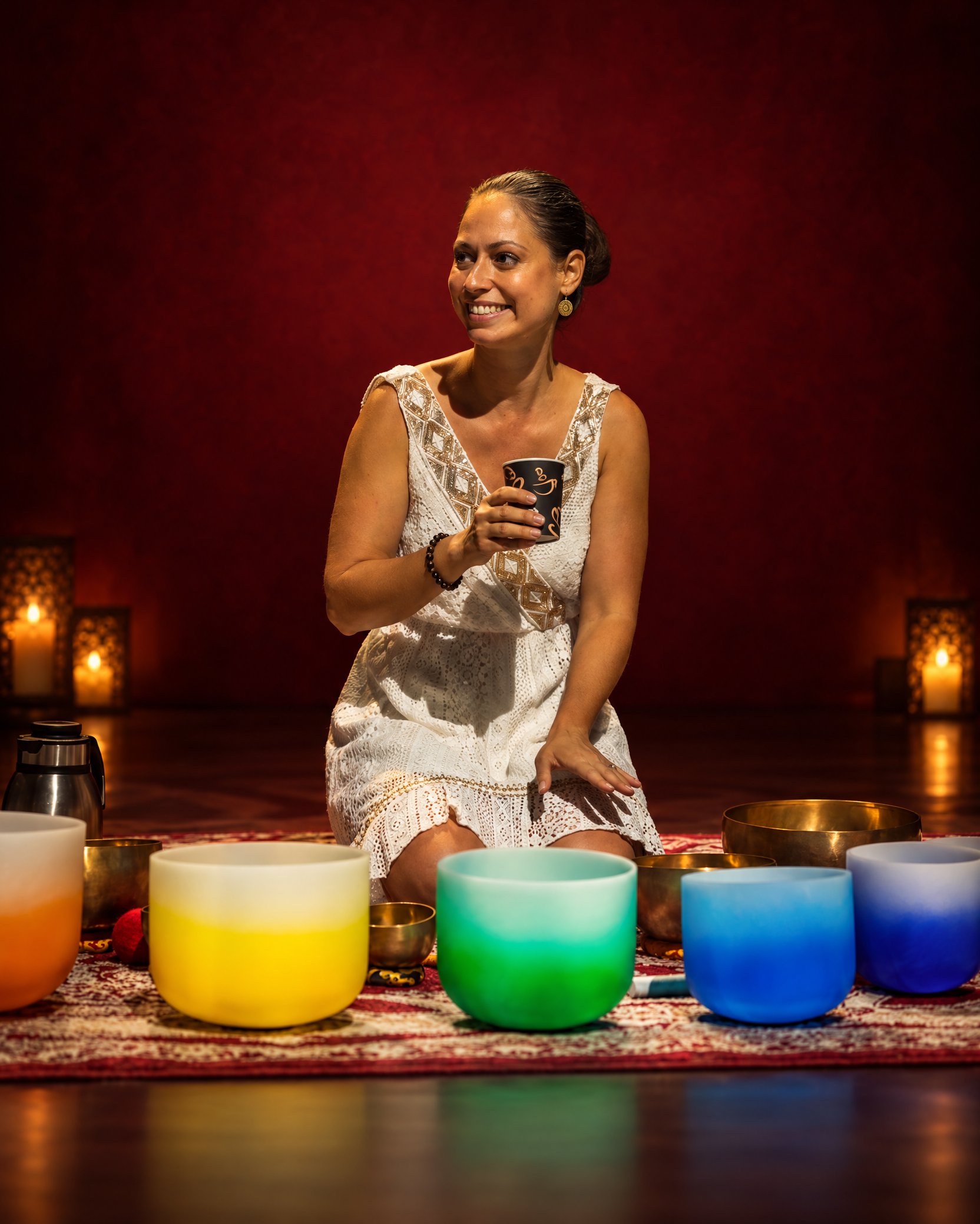 Szilvia holding ceremonial cacao among sound bowls against a deep crimson backdrop